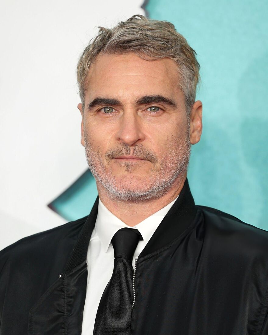 Close-up of a Joaquin Phoenix’s with green eyes, gray hair, and facial stubble wearing a black jacket and tie at an event. Close-up of a Joaquin Phoenix’s with green eyes, gray hair, and facial stubble wearing a black jacket and tie at an event.