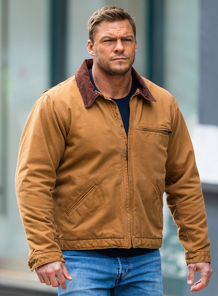 Alan Ritchson, Reacher star, wearing brown jacket and blue jeans, walking outdoors with a serious expression.
