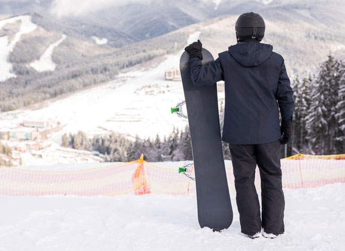 Person in winter gear holding a snowboard, overlooking snowy mountains, symbolizing harmless secrets people keep from partners.