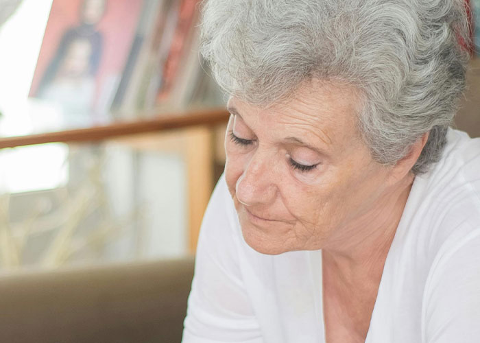 Elderly woman with gray hair looking down thoughtfully, reflecting on messy love triangle and chaotic secrets.