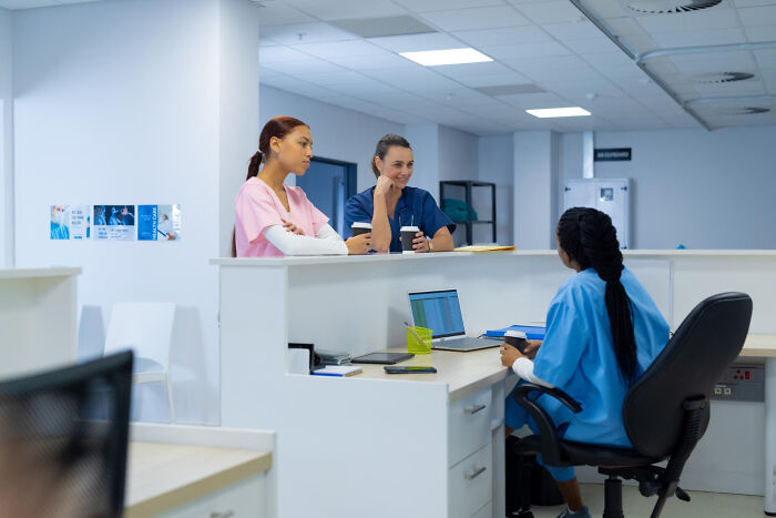 Three healthcare workers in scrubs having a discussion in a modern hospital nursing station environment