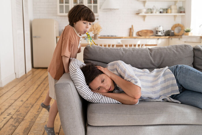 Tired woman resting on couch while child blows a party blower, illustrating common struggles women face not just part of being a woman.