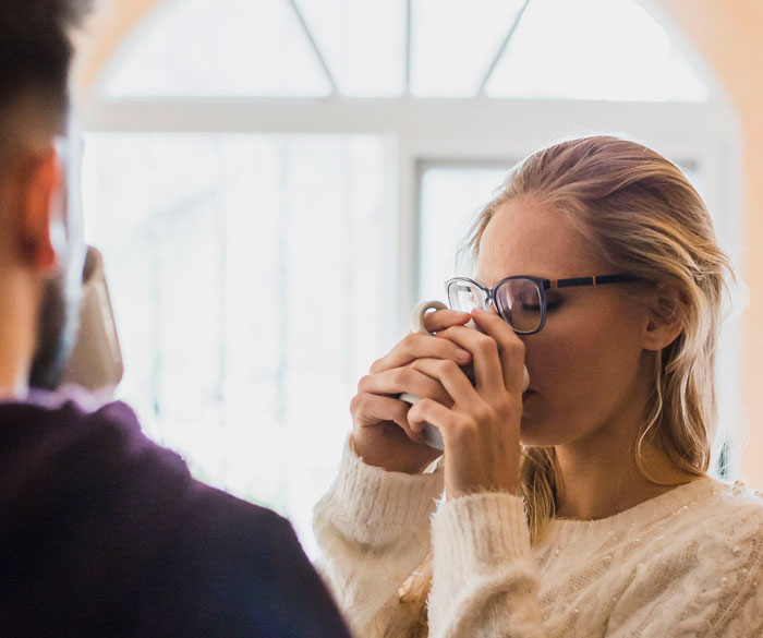 Woman with glasses drinking from a mug looking distressed after cutting off parents following betrayal with her fiancé.