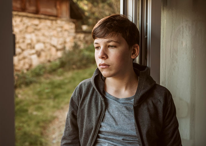Teen boy looking out window with a pensive expression, symbolizing grandparents guilt trip kid and family tension.