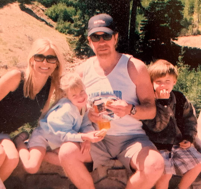 Os filhos de Chuck Norris sentados ao ar livre com o pai, aproveitando um dia de sol rodeado de natureza e vegetação.