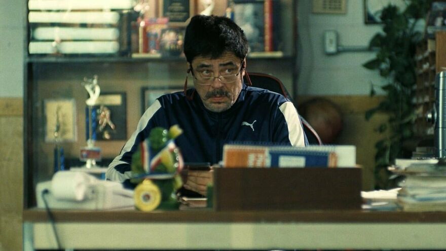 Man in a Puma jacket and glasses sitting at a desk surrounded by trophies and books, highlighting supporting performances.