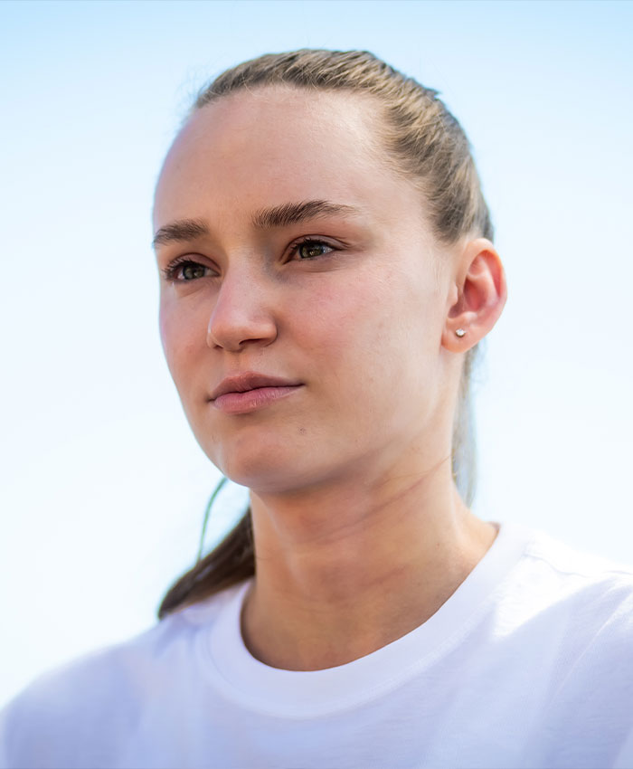 Elena Rybakina in a white shirt, looking serious outdoors, during a moment at a tennis trophy ceremony.