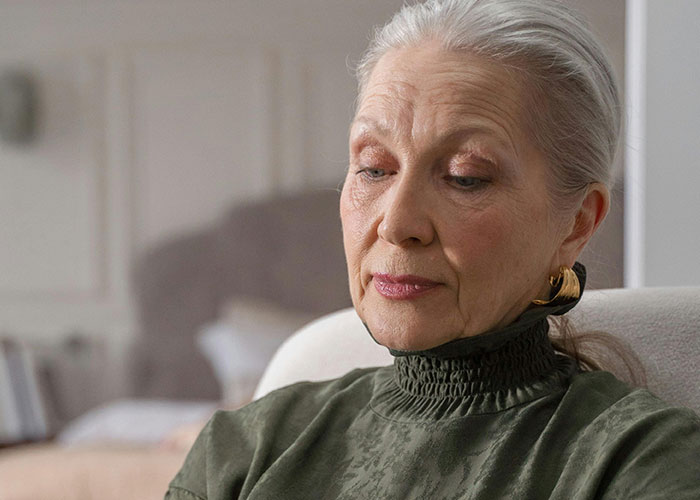 Elderly woman with silver hair and gold earrings, wearing a green blouse, appearing thoughtful in a cozy indoor setting.