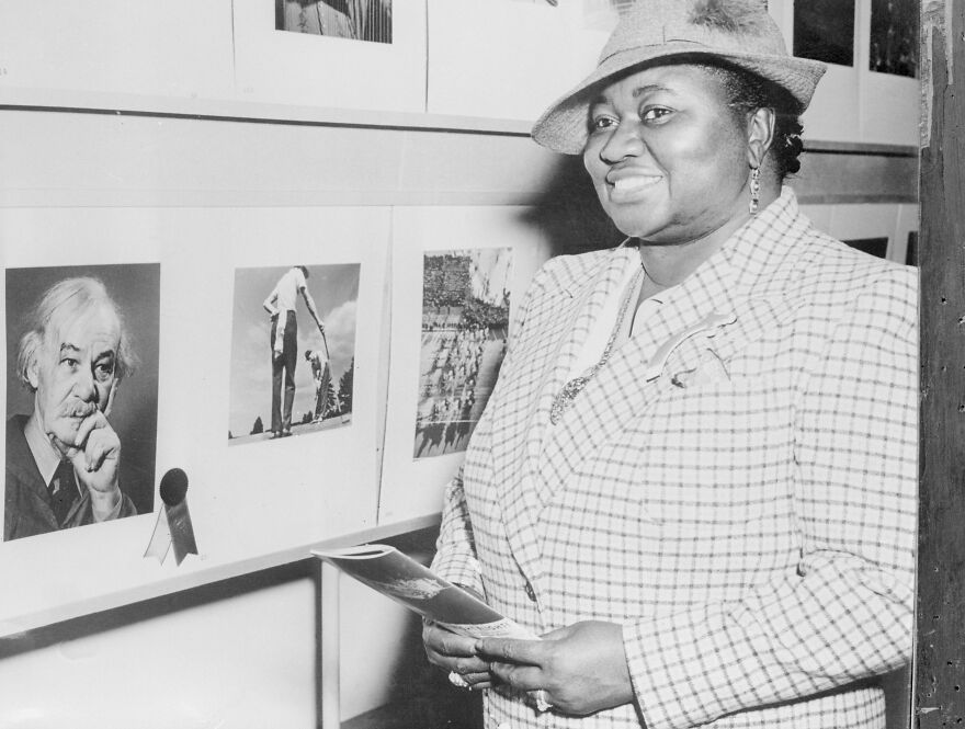 Hattie McDaniel sorrindo em uma exposição de arte, usando chapéu e casaco xadrez, representando a primeira negra a ganhar um Oscar.