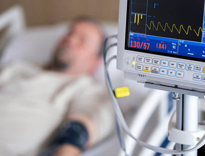 Hospital patient lying in bed with vital signs monitor showing blood pressure and heart rate readings, highlighting corporate secrets.