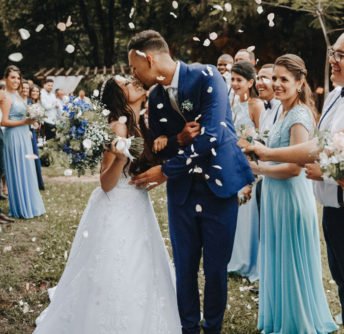 Bride and groom kissing outdoors surrounded by friends throwing flower petals at a joyful wedding celebration.