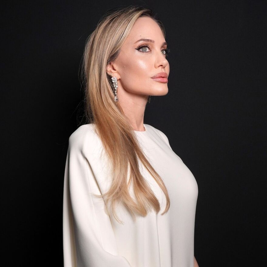 Angelina Jolie with long blonde hair and striking green eyes wearing elegant white dress and sparkly earrings against black background Angelina Jolie with long blonde hair and striking green eyes wearing elegant white dress and sparkly earrings against black background