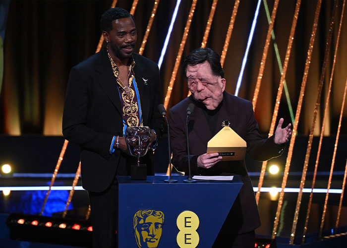 Adam Pearson ao lado de um homem em um evento do BAFTA, destacando os fãs gritando o tweet ofensivo de um comediante.