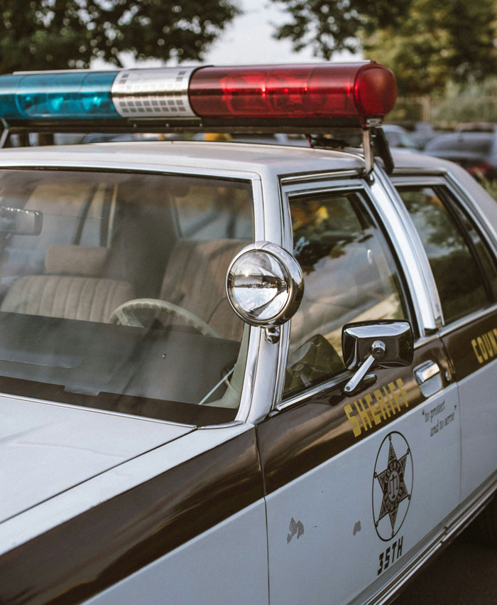 Police car with sheriff markings and red and blue emergency lights parked outdoors near trees.