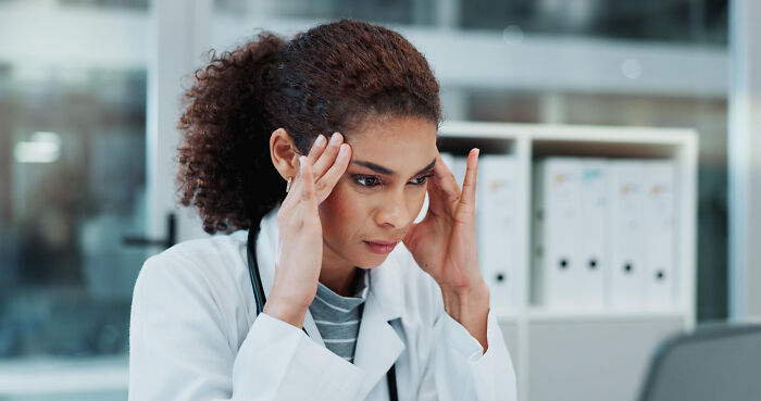 Doctor in white coat, stressed and holding temples, sitting at desk in modern medical office with files behind.