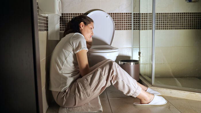 Woman experiencing discomfort sitting on a bathroom floor near toilet, illustrating period wardrobe and bathroom support group insights.
