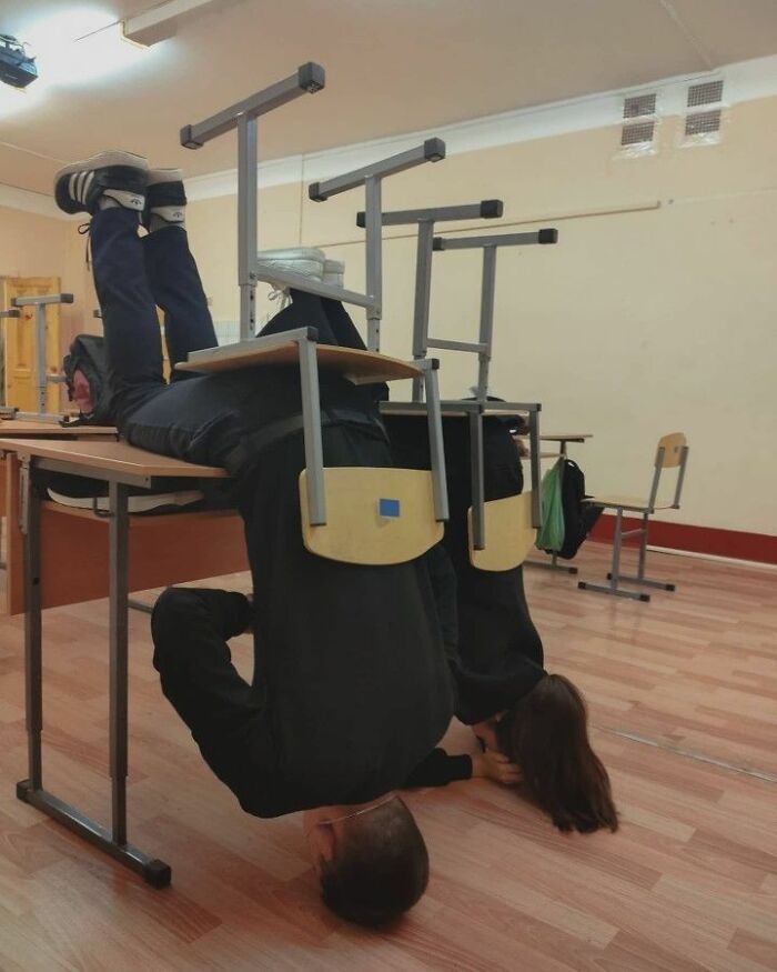 Two people pulling off creative poses by hanging upside down from school desks in a classroom setting.