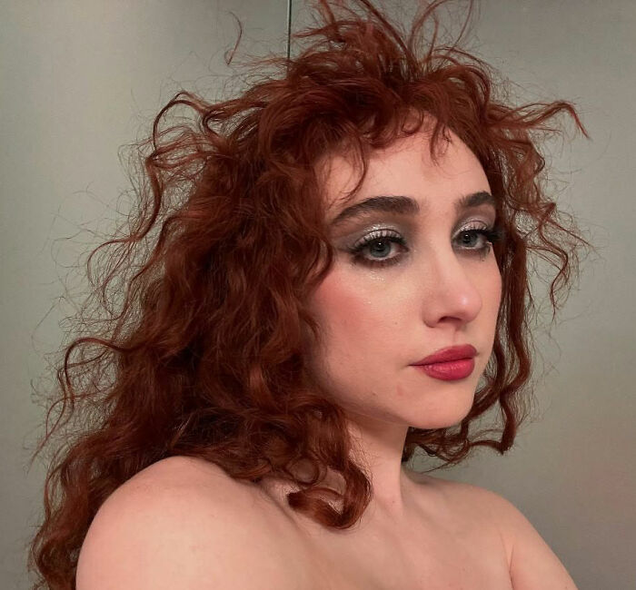 Woman with curly red hair and dramatic eye makeup, representing Chappell Roan in a contemplative pose indoors.
