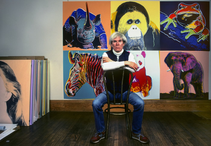 Andy Warhol sitting in front of colorful animal paintings, illustrating iconic imagery linked to famous leaders who changed history.