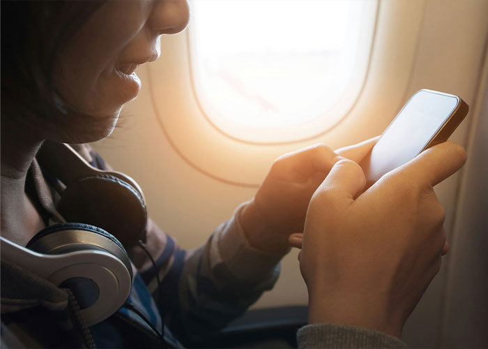 Passenger using phone near airplane window with headphones around neck on a major airline flight following new rule.