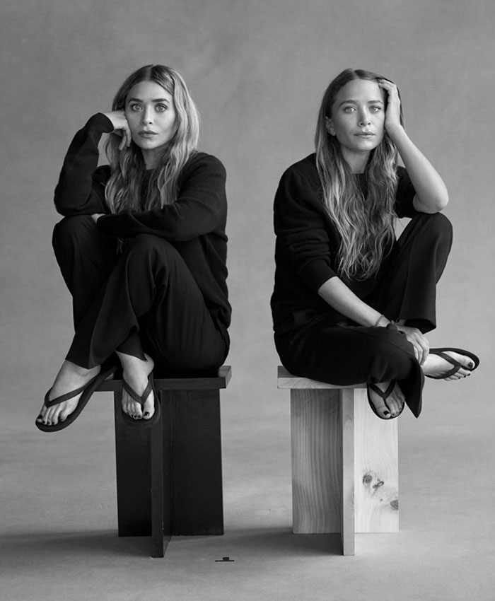 Mary-Kate and Ashley Olsen sitting on stools in black outfits, highlighting their disappearance and surprising rift.