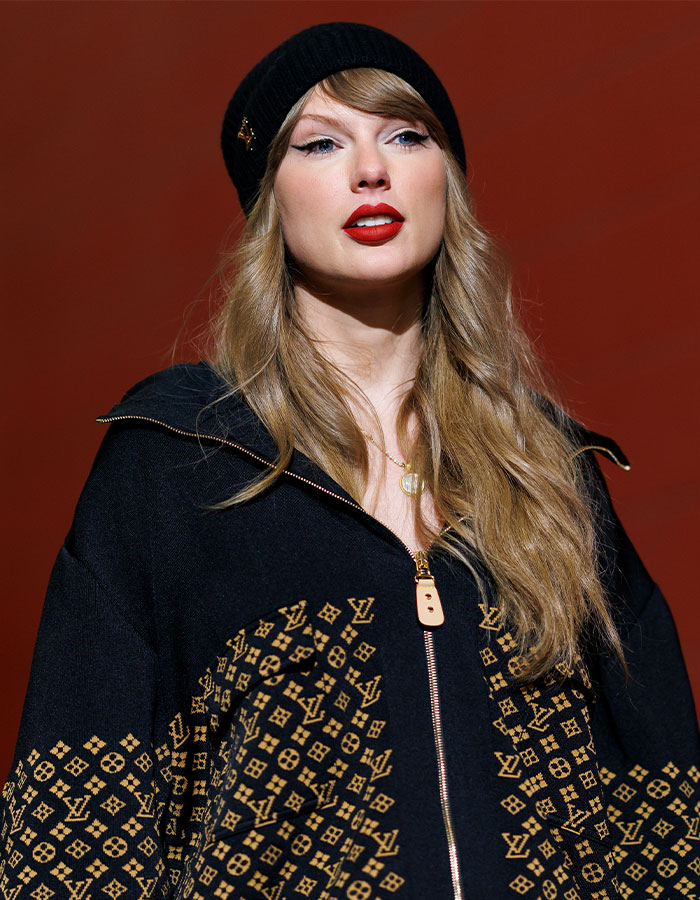 Taylor Swift wearing black beanie and Louis Vuitton jacket against a red background in a studio portrait.