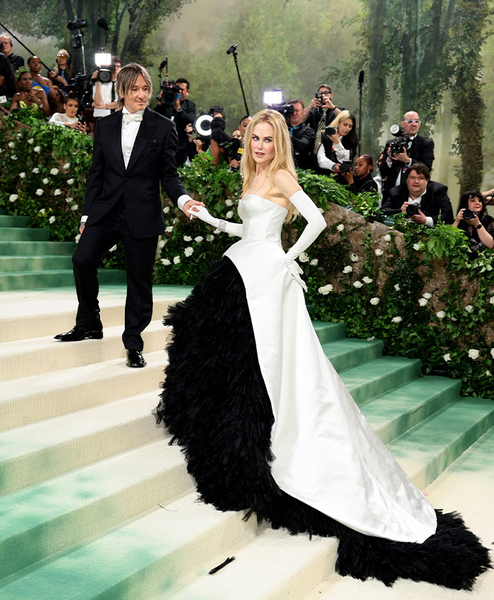 Nicole Kidman in black and white gown on staircase at event, surrounded by photographers and greenery background.
