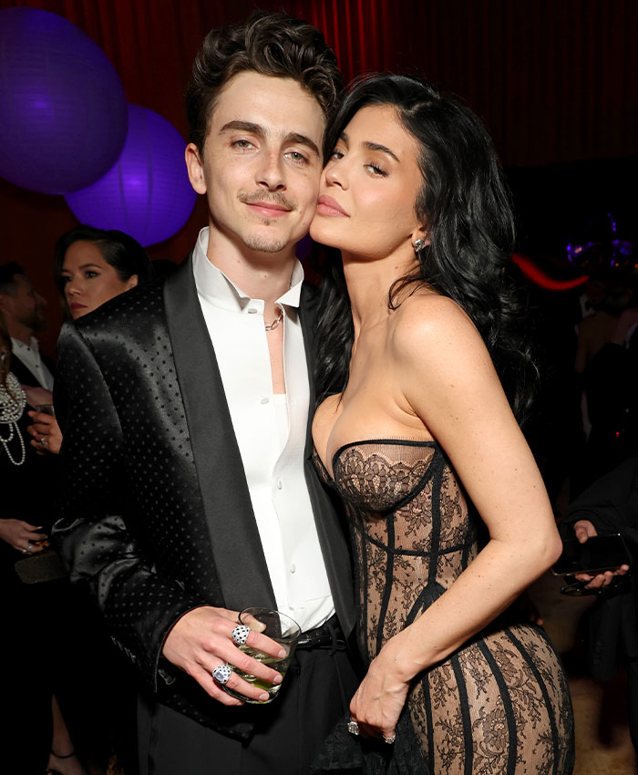 Timoth&eacute;e Chalamet at an event with a guest, dressed in a black tuxedo jacket and white shirt, posing for a photo.