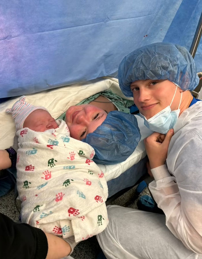Newborn swaddled in hospital blanket with two young family members wearing surgical caps after birth.