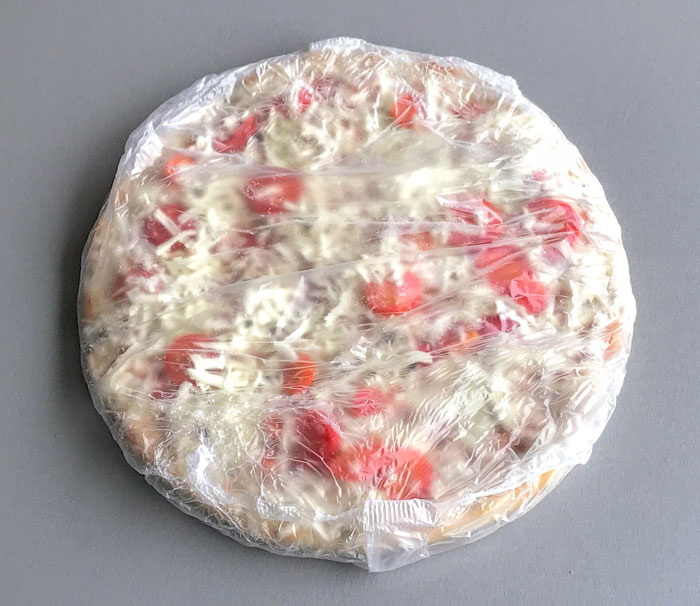 Frozen pizza topped with cheese and red peppers wrapped in clear plastic wrap on a gray surface.