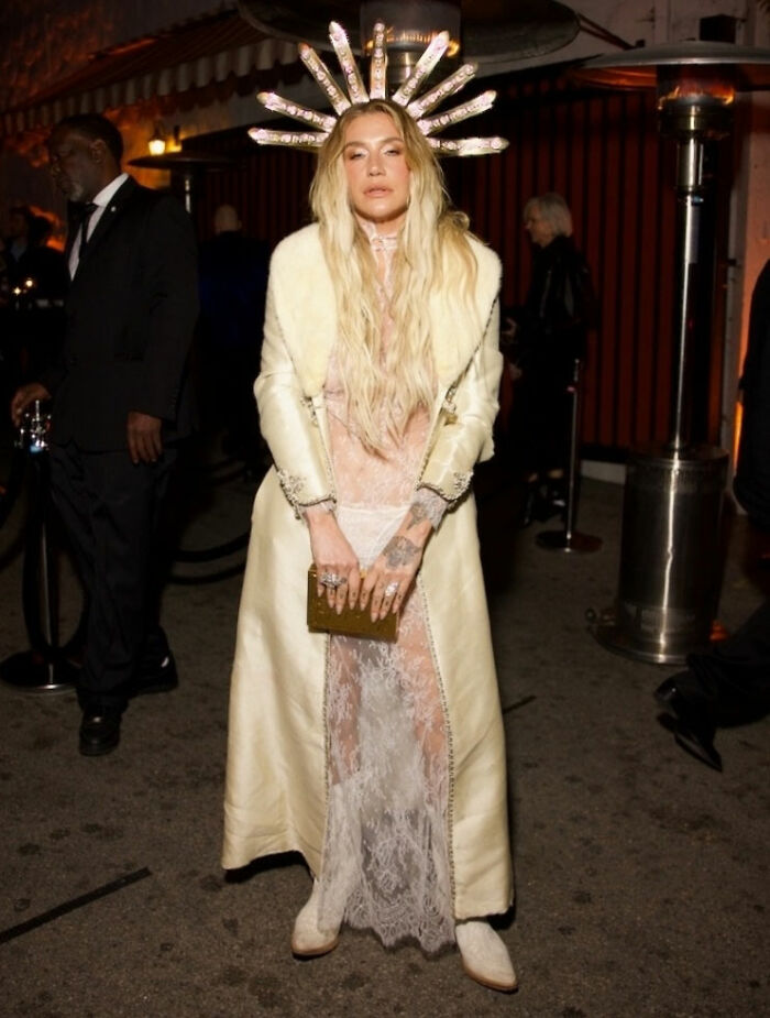Woman in a long coat and sheer lace dress with an elaborate headpiece at the 2026 award season fashion fails event.