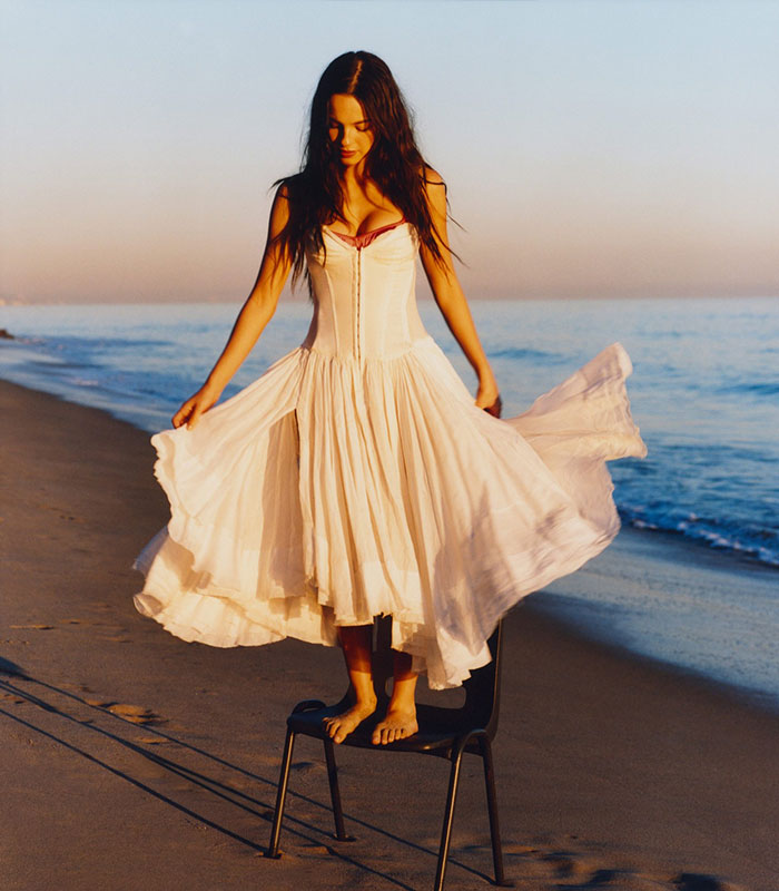 Olivia Rodrigo in a flowing white dress standing barefoot on a chair by the beach during sunset for Vogue cover. Olivia Rodrigo in a flowing white dress standing barefoot on a chair by the beach during sunset for Vogue cover.