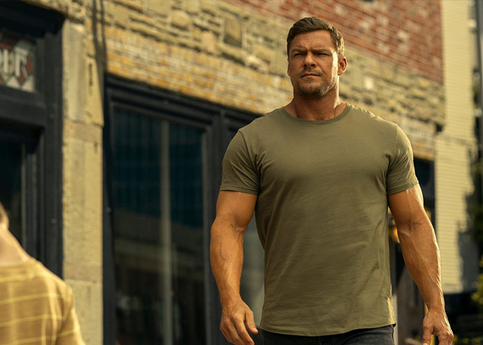 Reacher star Alan Ritchson walking outside a building in a green t-shirt with a serious expression on his face.