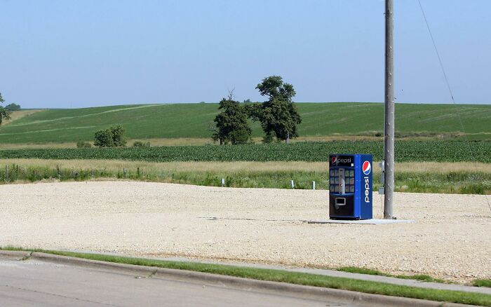 Pepsi vending machine standing alone on a gravel lot with green fields and clear sky in a US rural landscape.