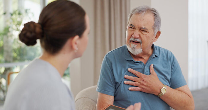 An elderly man in a blue shirt discussing health concerns with a female doctor in a bright home setting.