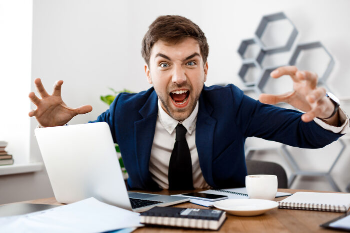 Angry man in suit at desk with laptop, aggressively reaching forward, illustrating karma hitting those who try to sabotage.