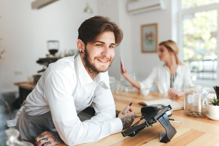 Young barista with tattoos smiling at cafe counter while a woman uses her phone in the background, discussing Gen Z stare reactions.