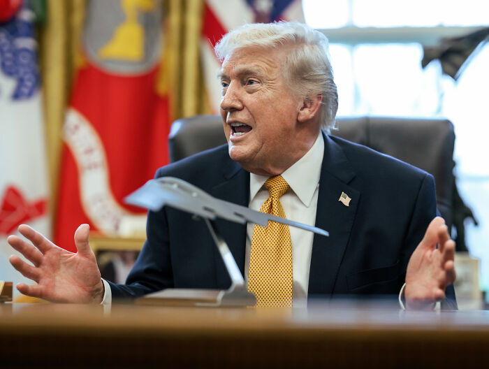Former President Donald Trump speaking in the Oval Office with a model airplane on the desk behind flags.
