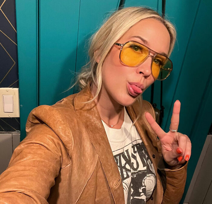 Anna Camp wearing yellow sunglasses and a brown jacket, playfully posing with a peace sign indoors.