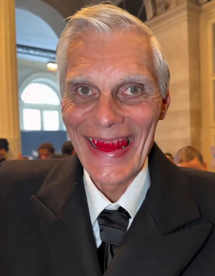 Elderly man at Paris fashion week wearing unique makeup and a formal black suit with fans comparing looks to Hunger Games.