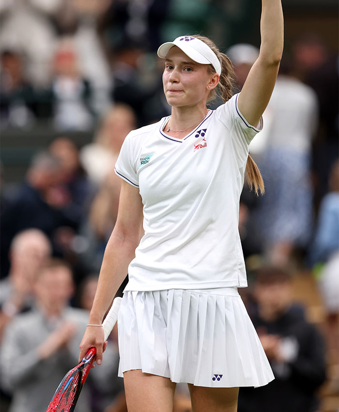 Tennis star Elena Rybakina in white outfit holding racket and raising hand during trophy ceremony on court.