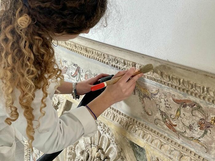Woman in a lab coat carefully restoring a decorative wall panel using specialized brushes and tools for detailed work.