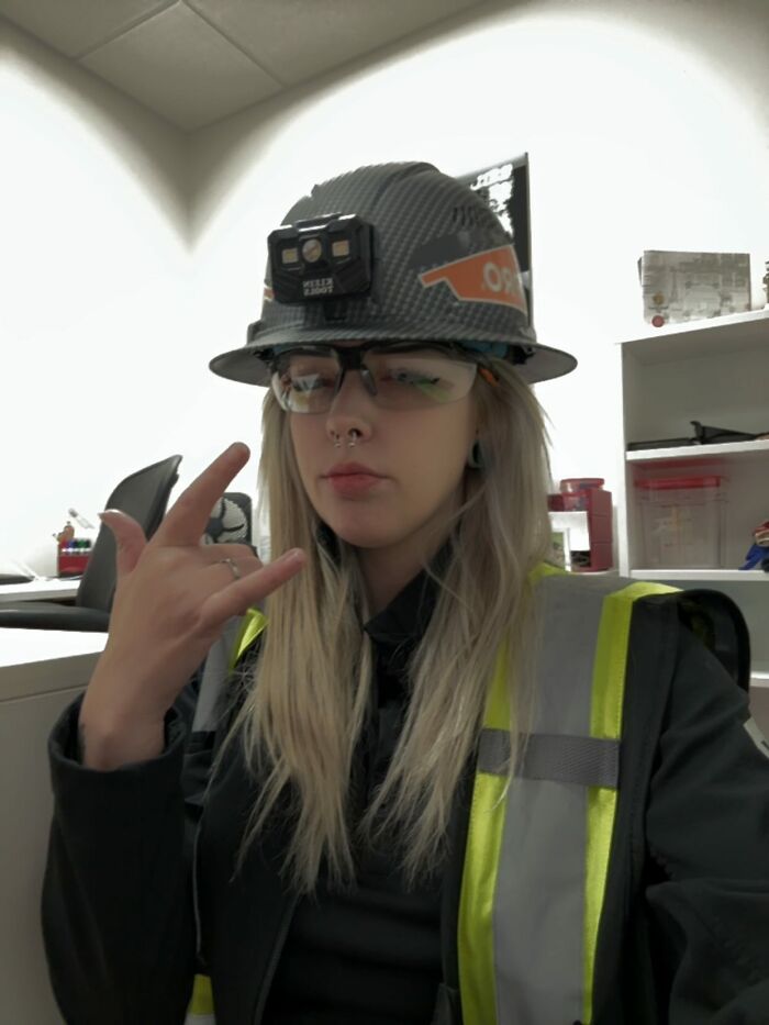 Woman wearing a hard hat and safety glasses, showcasing power tools and lab coats in an industrial workplace setting.