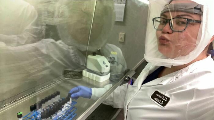 Woman in a lab coat and protective gear working with test tubes and power tools in a scientific setting.