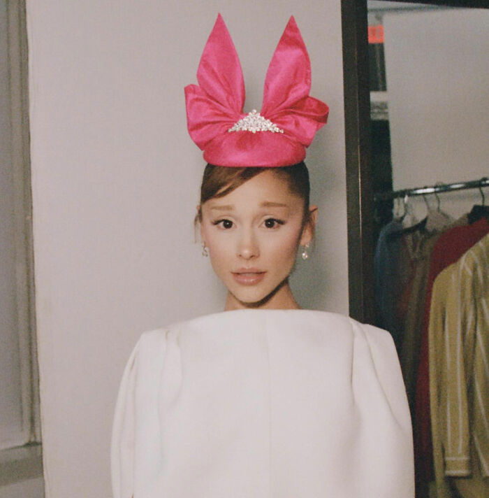 Ariana Grande wearing a bright pink headpiece and white outfit, sparking plastic surgery rumors with her latest look.