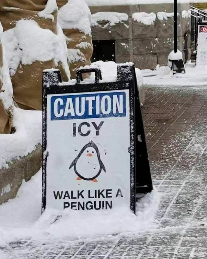 Snow-covered caution sign warning icy conditions with a funny penguin reminding people to walk carefully.