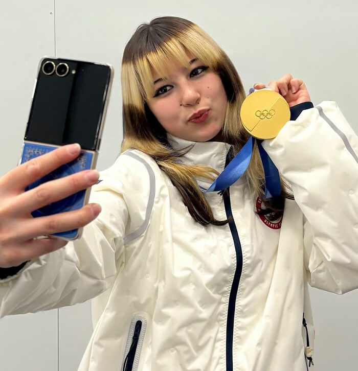 Alysa Liu taking a selfie holding an Olympic gold medal, wearing a white jacket with a blue ribbon.