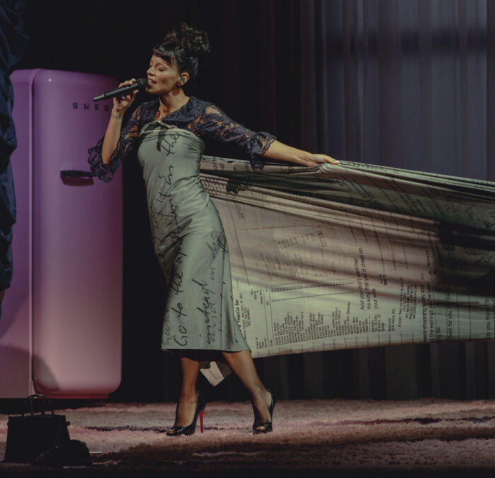 Woman singing on stage, wearing a printed dress and holding a flowing fabric, related to David Harbour’s alleged partner event.