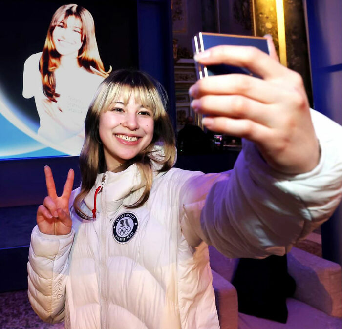 Olympic hero Alysa Liu in a white jacket taking a selfie while smiling and flashing a peace sign indoors.