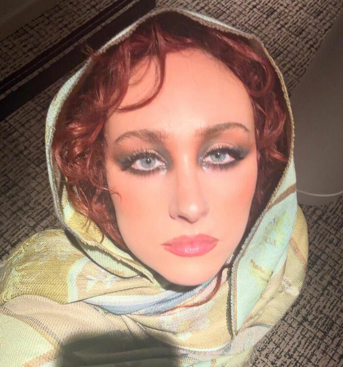 Chappell Roan wearing a patterned hooded shawl with dramatic eye makeup in a close-up selfie indoors.